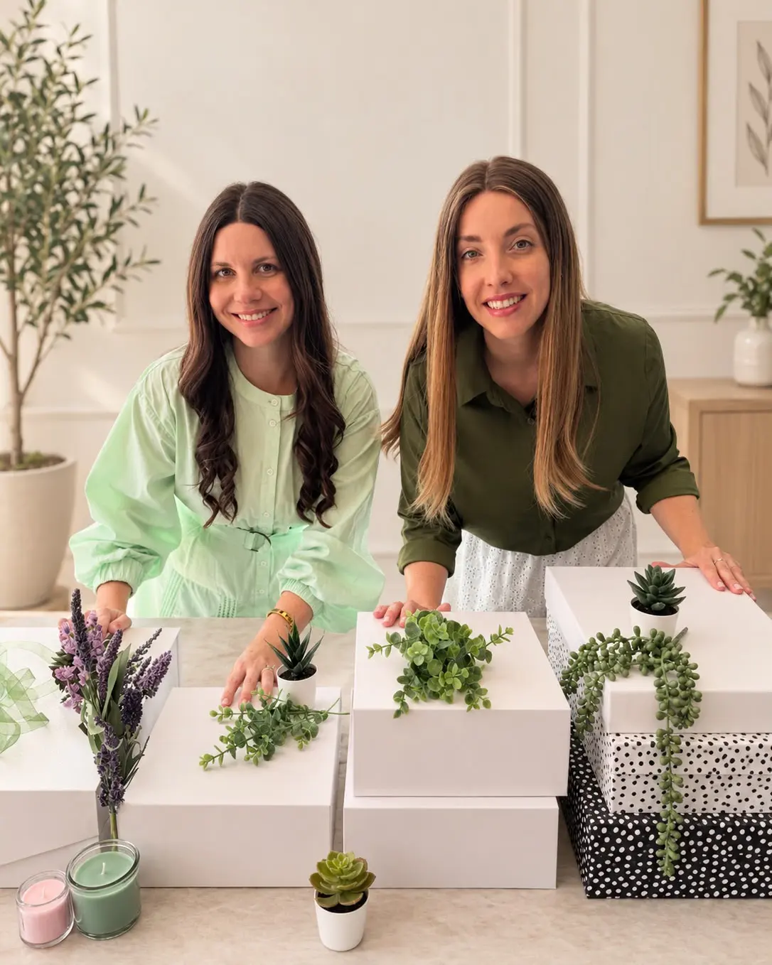 The two founders of Basket & Bond smiling behind a table of white and polka-dot gift boxes they are assembling, with plants and candles.